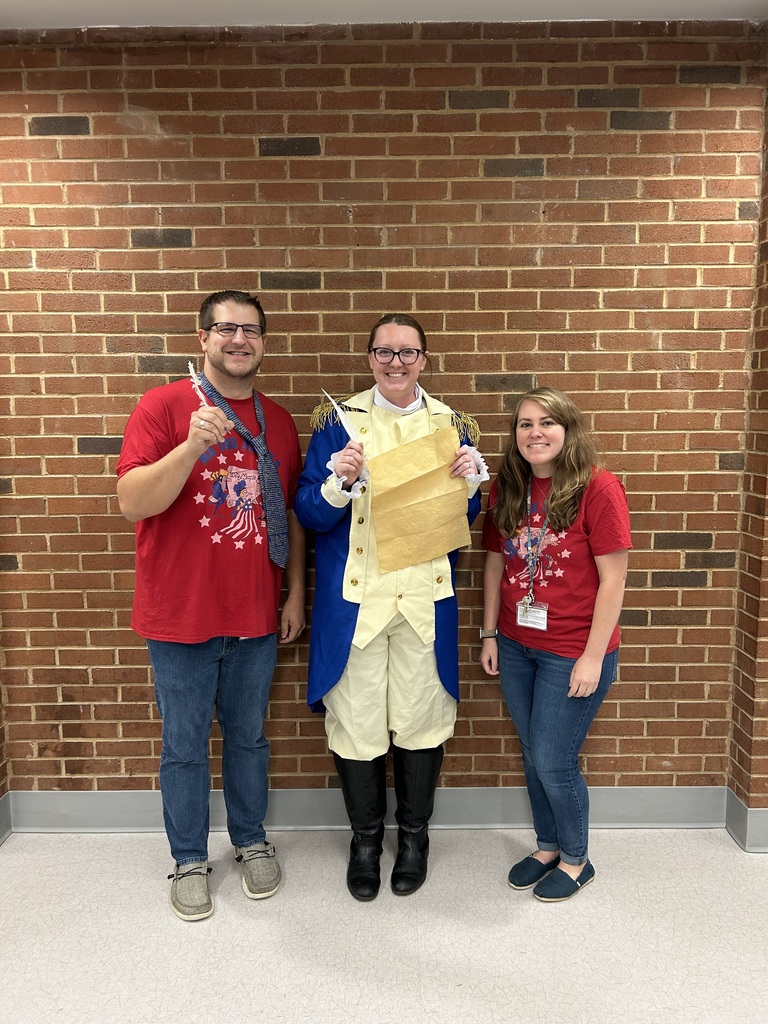 Three CMS staff members pose in front of a brick wall during a Constitution Day celebration. The person in the center is dressed in a colonial-style blue, cream, and gold costume and holds a quill pen and parchment, while the two staff members beside them wear matching red patriotic shirts.