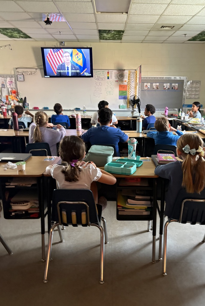 A classroom with students seated at desks, a television mounted on the wall, and a whiteboard with papers attached.
