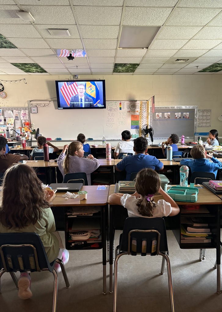 A classroom with students seated at tables, watching a presentation on a mounted TV screen. Desks have books, water bottles, and trays.
