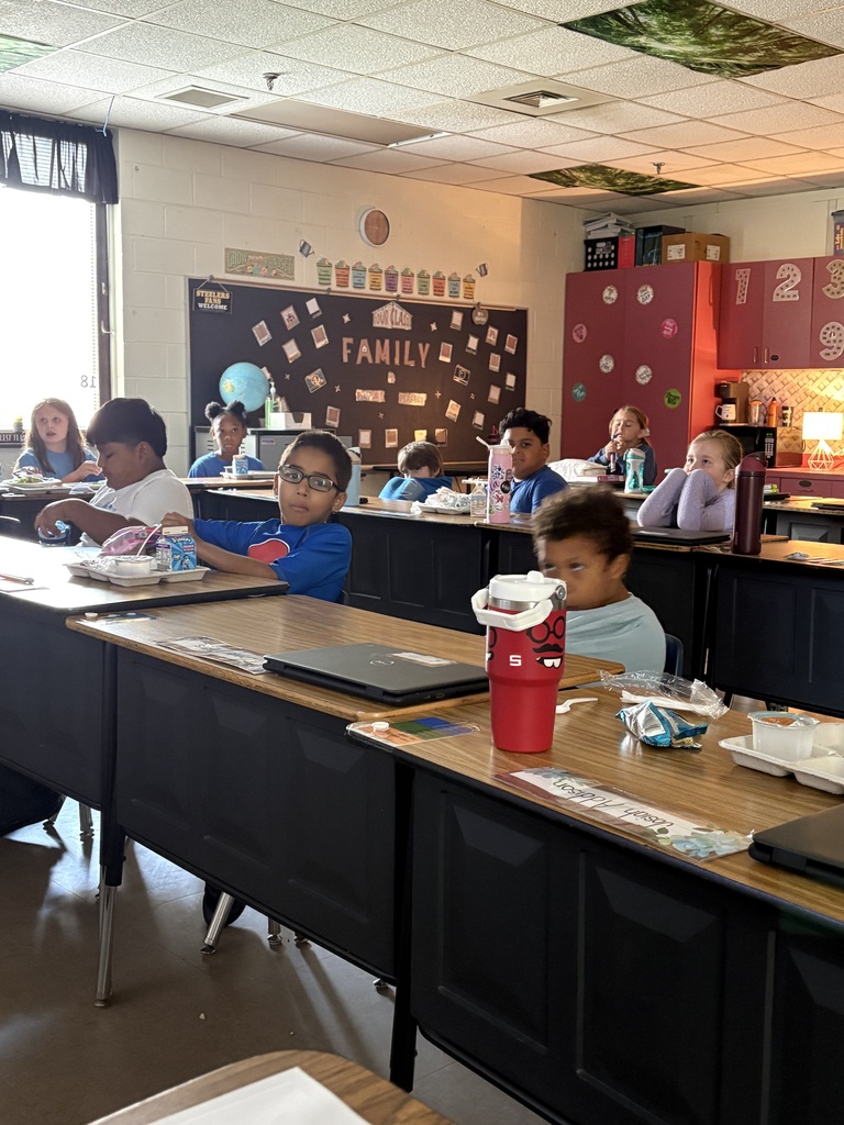 A classroom with students seated at tables, one using a laptop. A red cup is on the table.