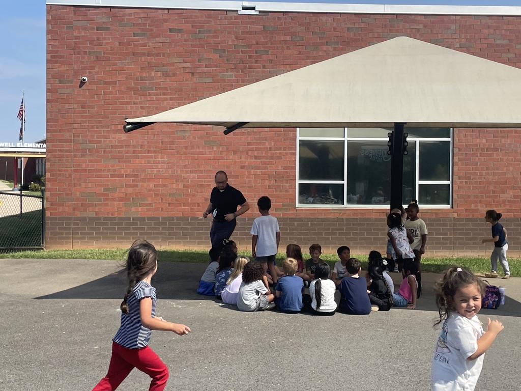 A group of children in front of a brick building. An adult is standing in front of them.