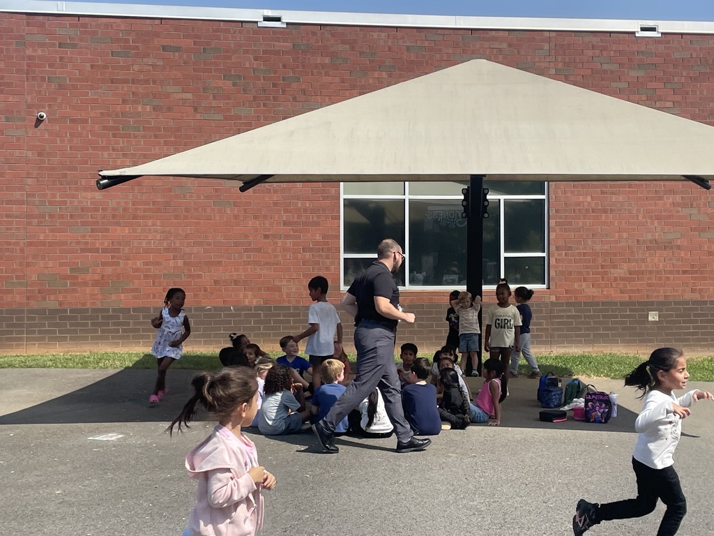 A group of children play outdoors near a brick building. A man is walking through a crowd of children.