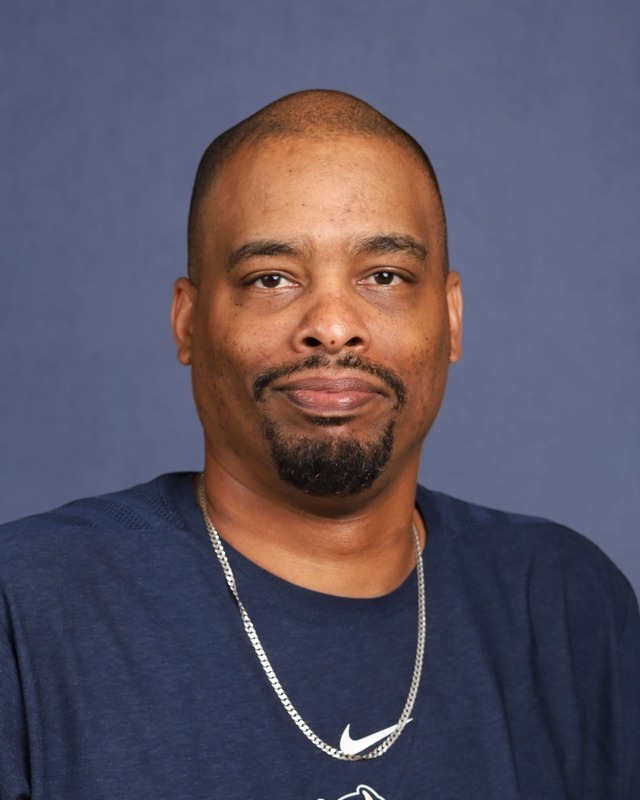 A man with a beard and mustache wearing a blue shirt and a silver Nike necklace poses against a blue background.