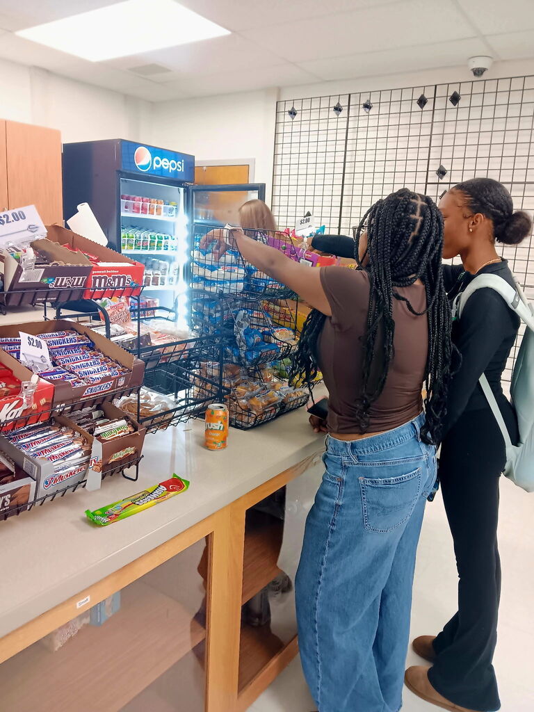 Two people near a food counter in a brightly lit room. Various snacks in containers and a Pepsi cooler.