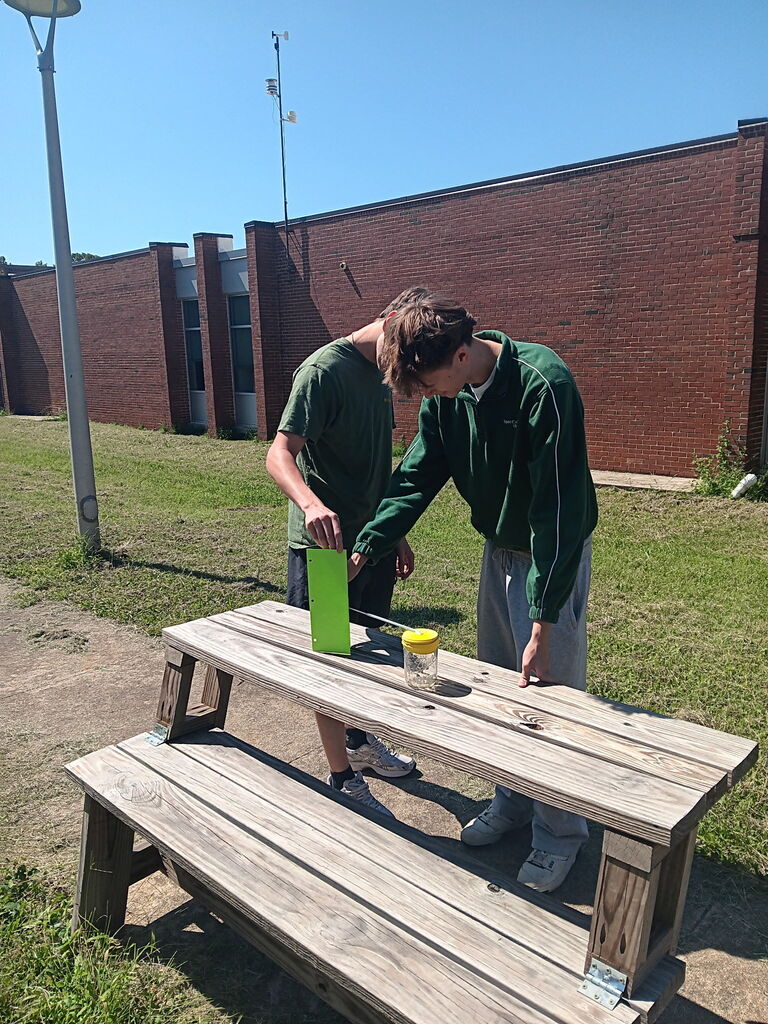 Two individuals examine a green item on a wooden table. They wear matching green shirts. Behind them is a brick building.