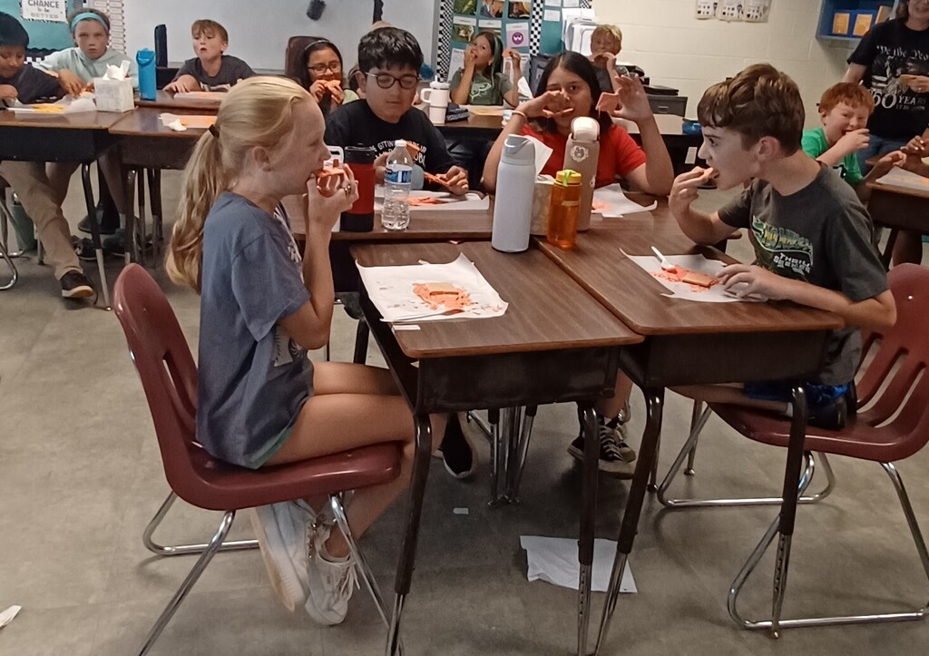 Classroom with students eating at desks. Two kids with food in hands. Some desks have bottles, papers.