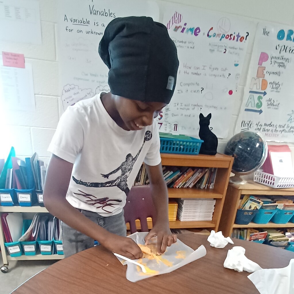 A child in a white shirt and black beanie is at a desk, using a knife on paper.