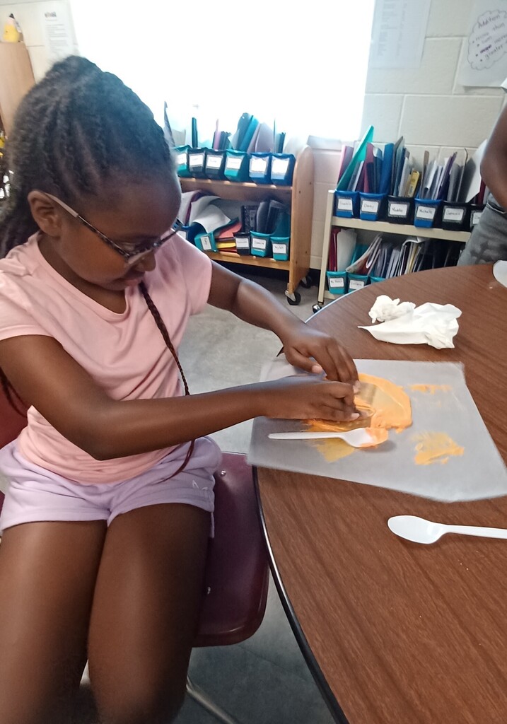 A young girl with glasses sits at a table, wearing a pink shirt. She holds a spoon and works on an art project.