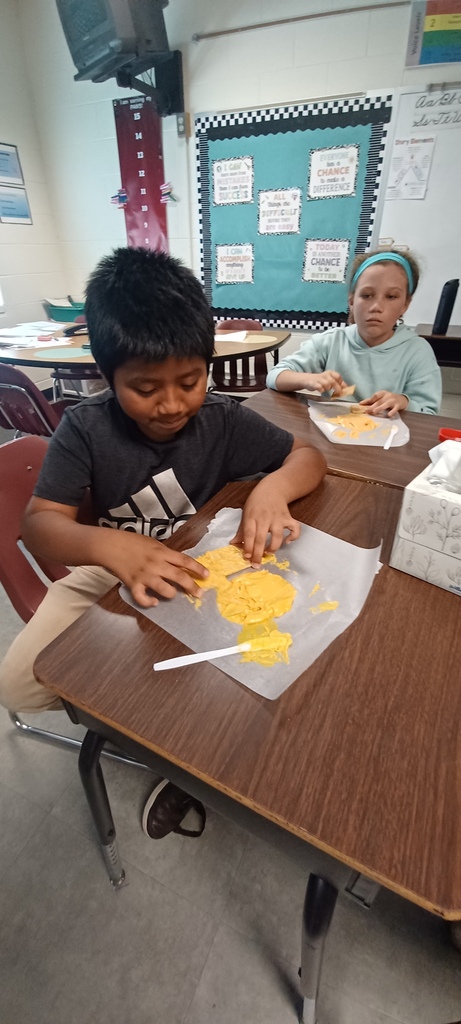 Two children sit at a desk, working on a project. One child is focused on a yellow substance on a paper.