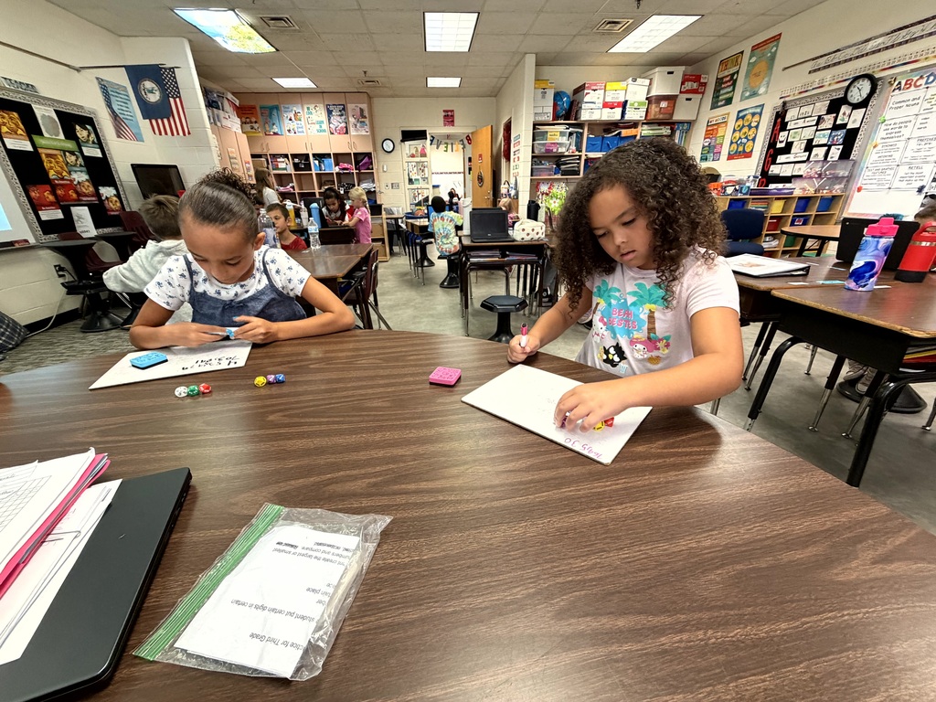 Two children at a desk in a classroom, each working on paper with markers. A flag, shelves, and other students are in the background.