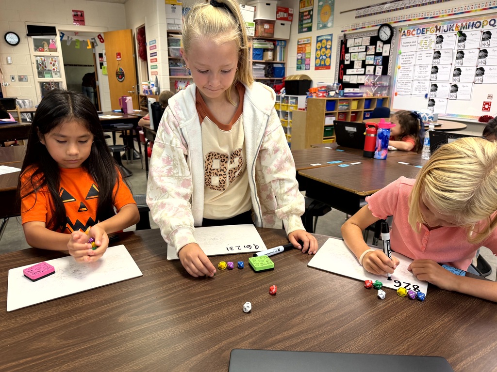 Three students in a classroom, seated at a table, engaged in a learning activity with paper and small objects.