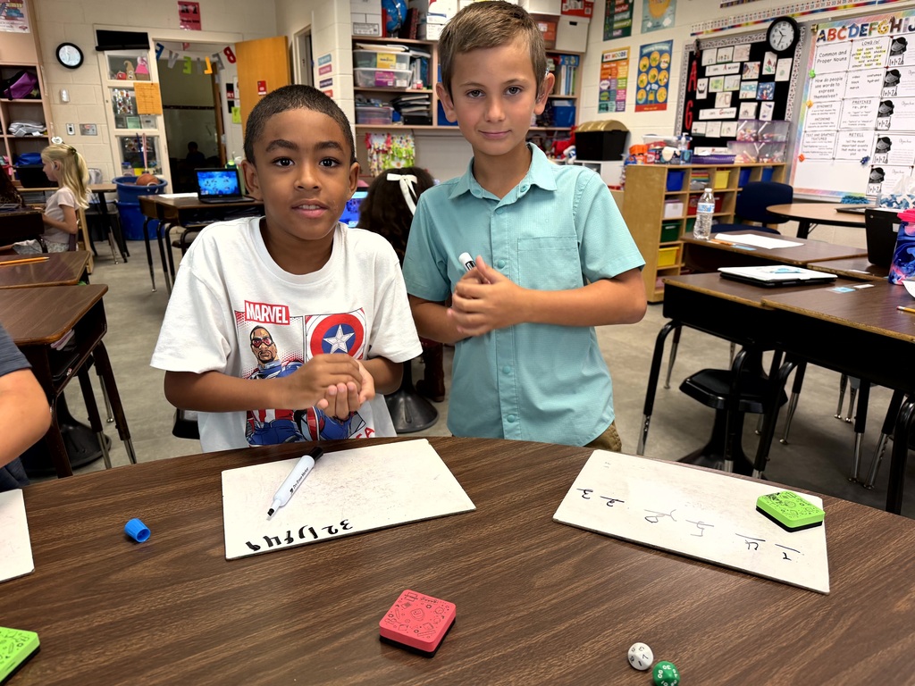 Two young boys in a classroom with desks behind them, one holding a pen and writing on paper.