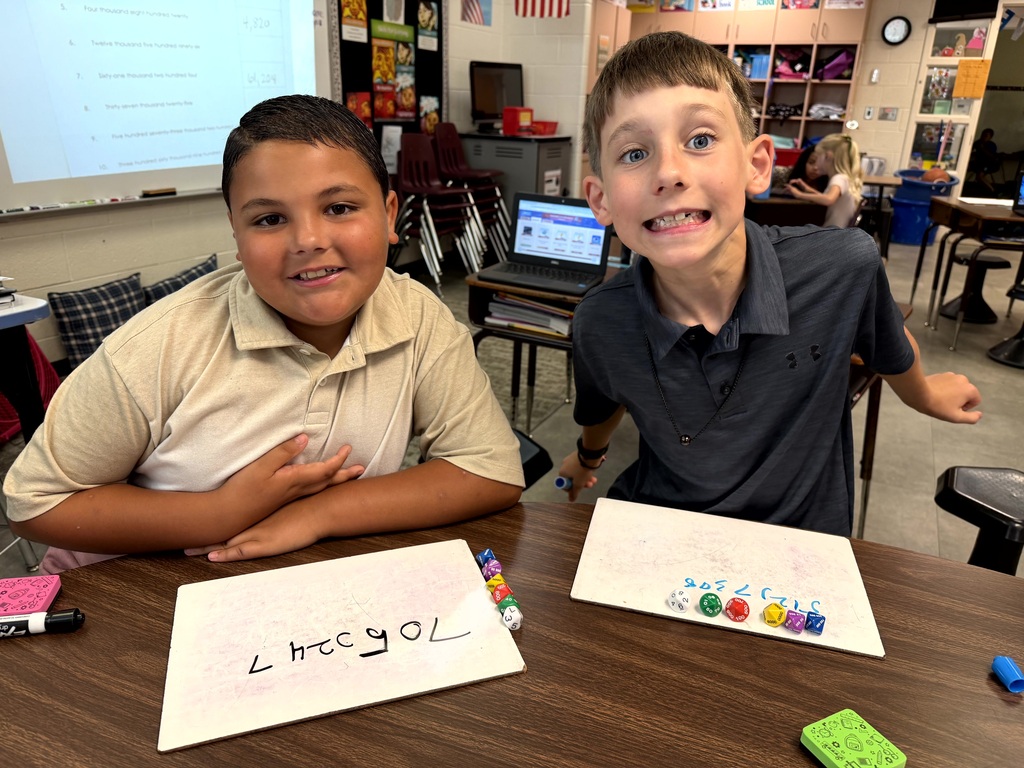 Two boys sit at a desk with papers. One has "LHCGYOL" on it, the other has numbers. A laptop and chairs are in the background.