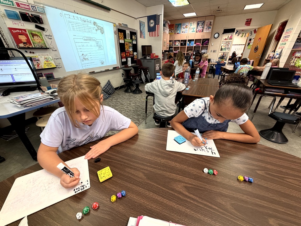 Two students focus on their work at a table in a classroom, with a whiteboard in the background.