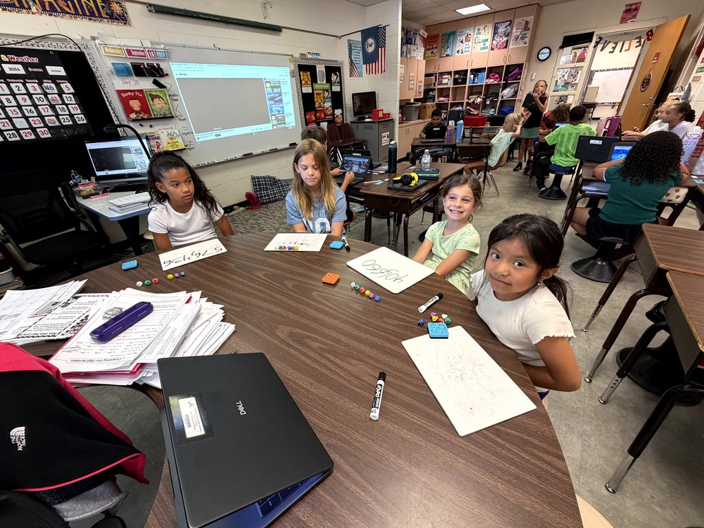 Young children sit at a desk in a classroom, working with papers and laptops. Other children are seated at desks around them.