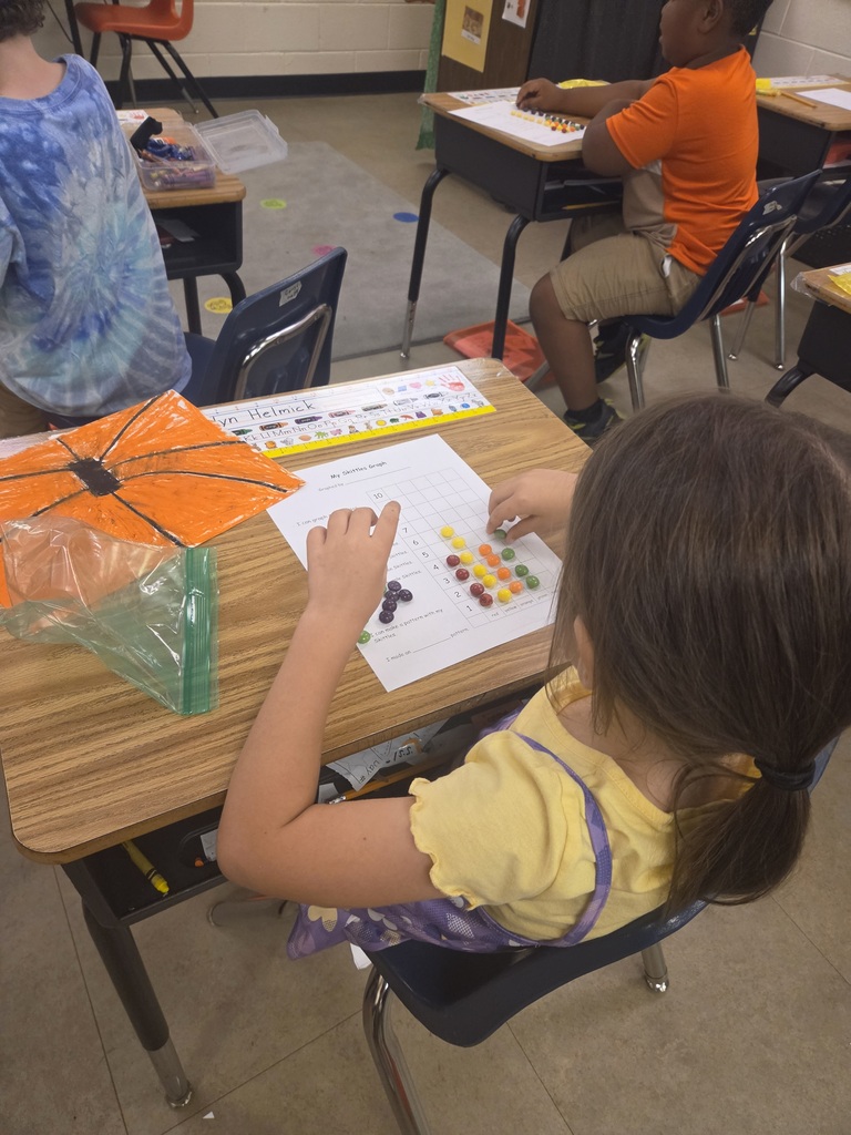 Children seated at desks in a classroom, one using colored beads on a paper with a pumpkin, another with a colorful tie-dye shirt.