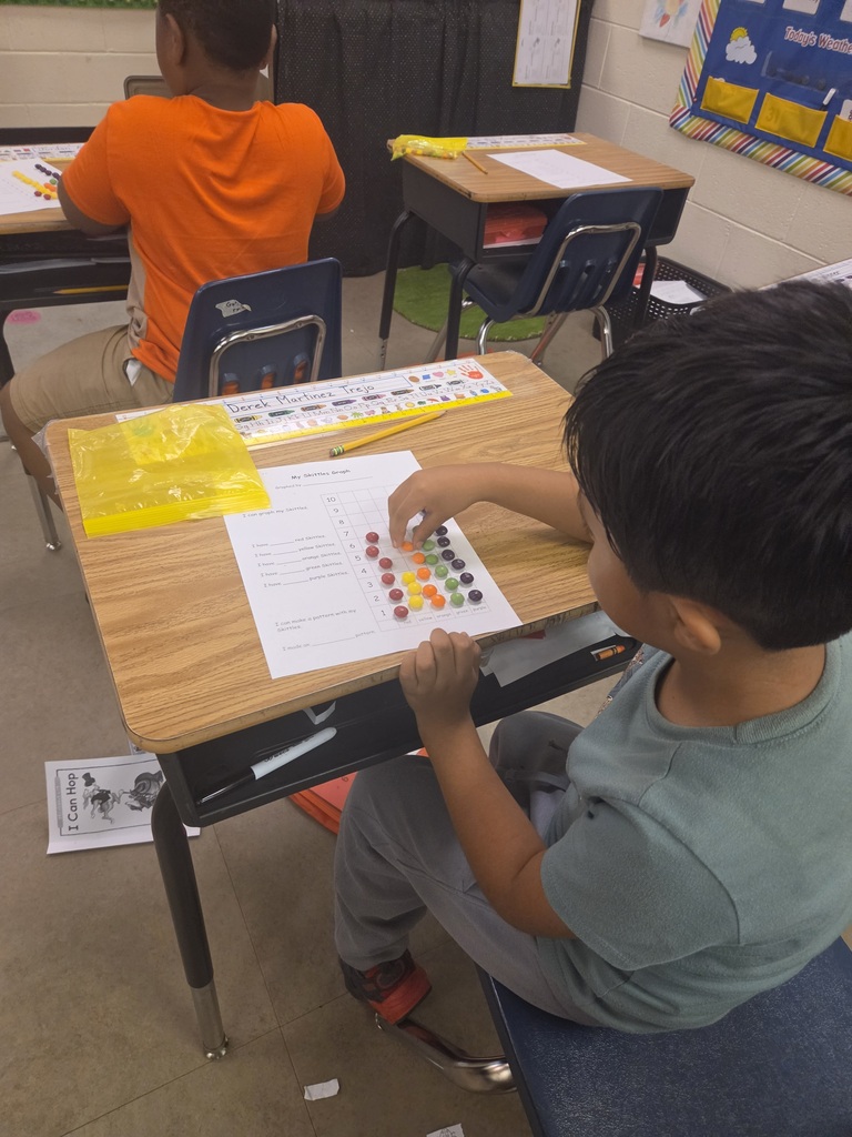 A child uses colorful beads to count numbers on a desk in a classroom. Another child sits in the background.