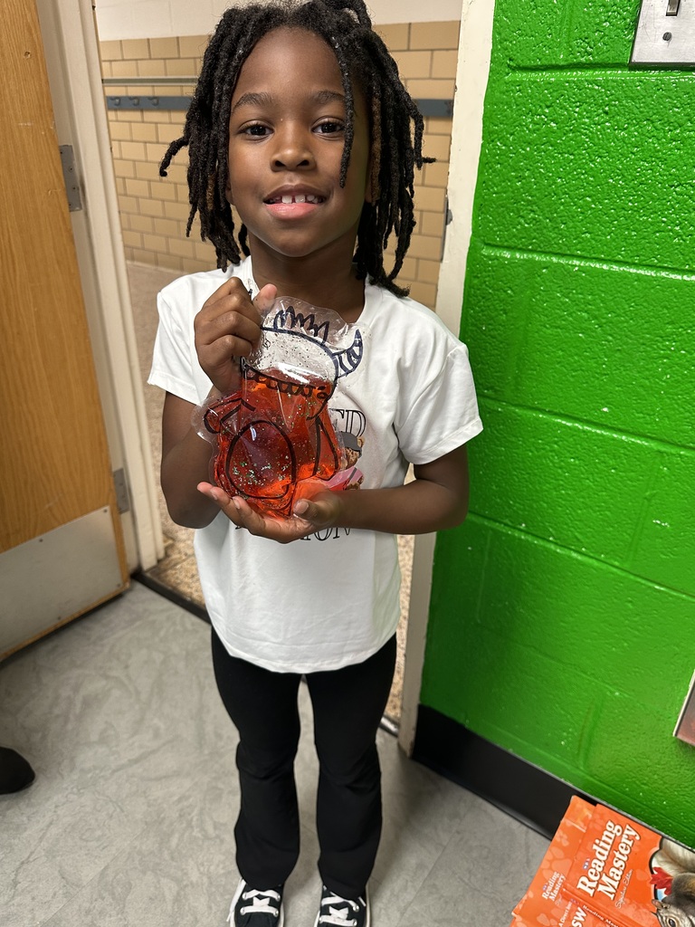 A young child holds an orange glass object and smiles, standing in front of a green wall.