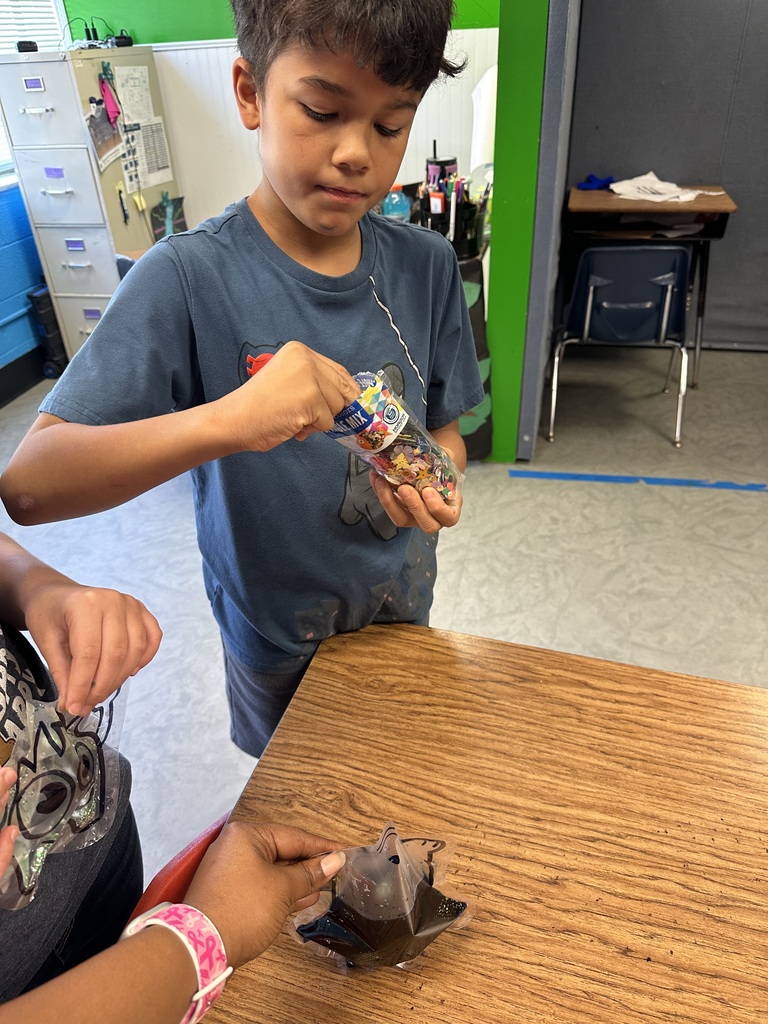 A boy holds a plastic bag filled with colored items in a classroom. Another child sits at a table nearby.