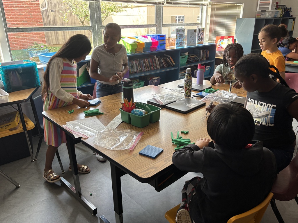 Kids sit at a table in a classroom. Some have crafts and supplies on the table.