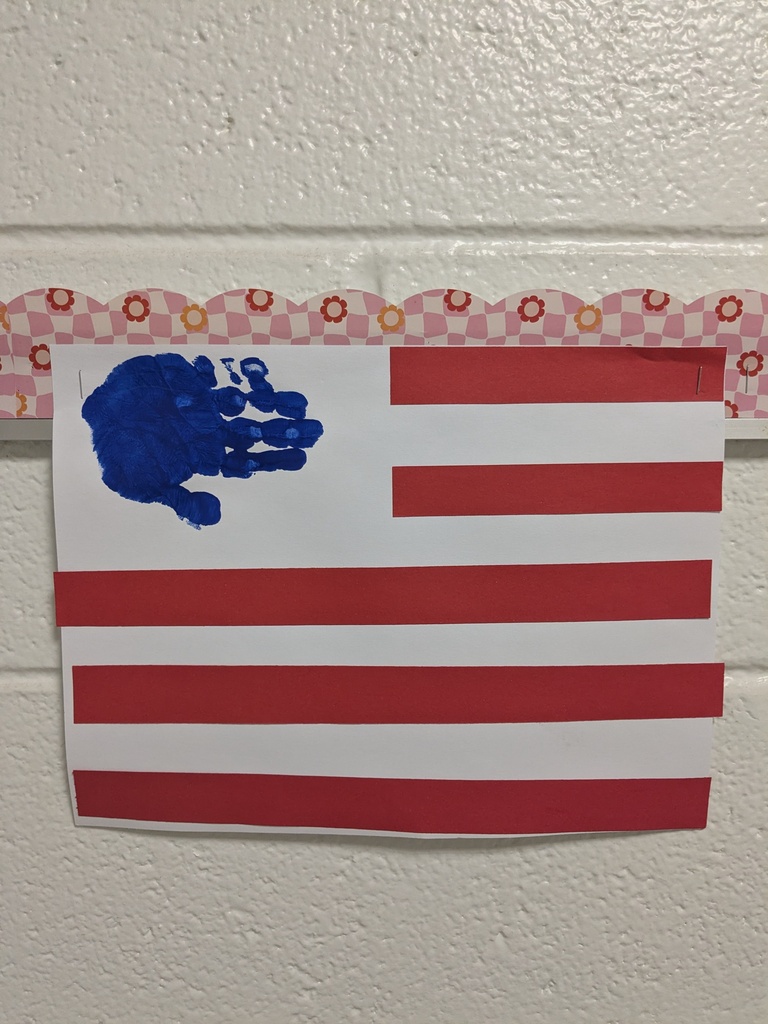 A handmade flag with 13 red and white stripes and a blue field with white stars is attached to a white brick wall.