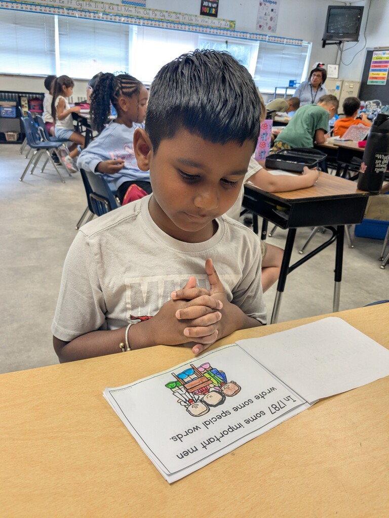 A child in a classroom, eyes closed, hands clasped together. A book with colorful pictures and text is in front of him.