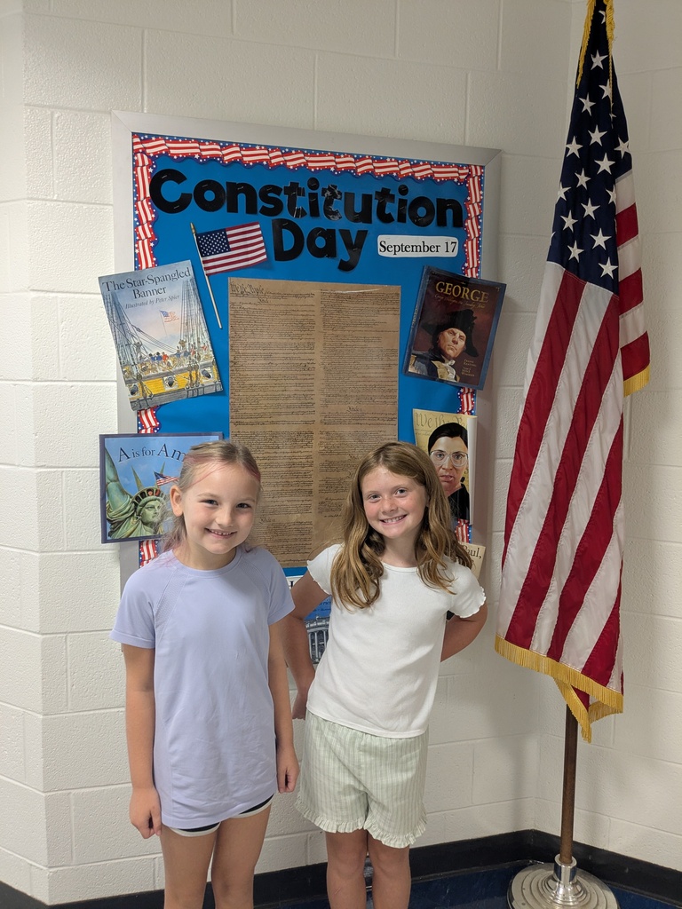 Two young girls stand next to an American flag in a classroom with a Constitution Day poster.