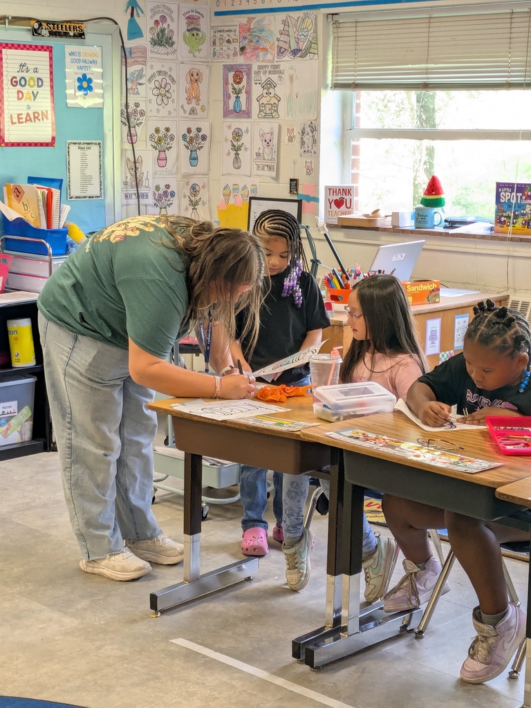 Adult and three children in a classroom, with a woman leaning over a table, and children sitting at desks.