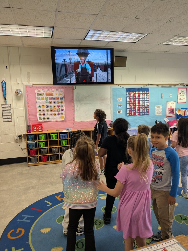 Classroom with children watching a television showing a man. Rug, shelves with bins, and colorful walls.