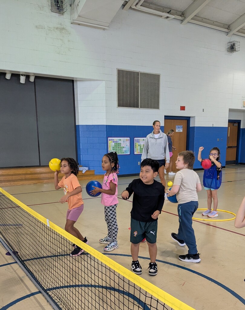 Students stand on either side of a low net while practicing throwing, catching, and tossing colorful playground balls. Teachers supervise and give instructions as students take turns participating.