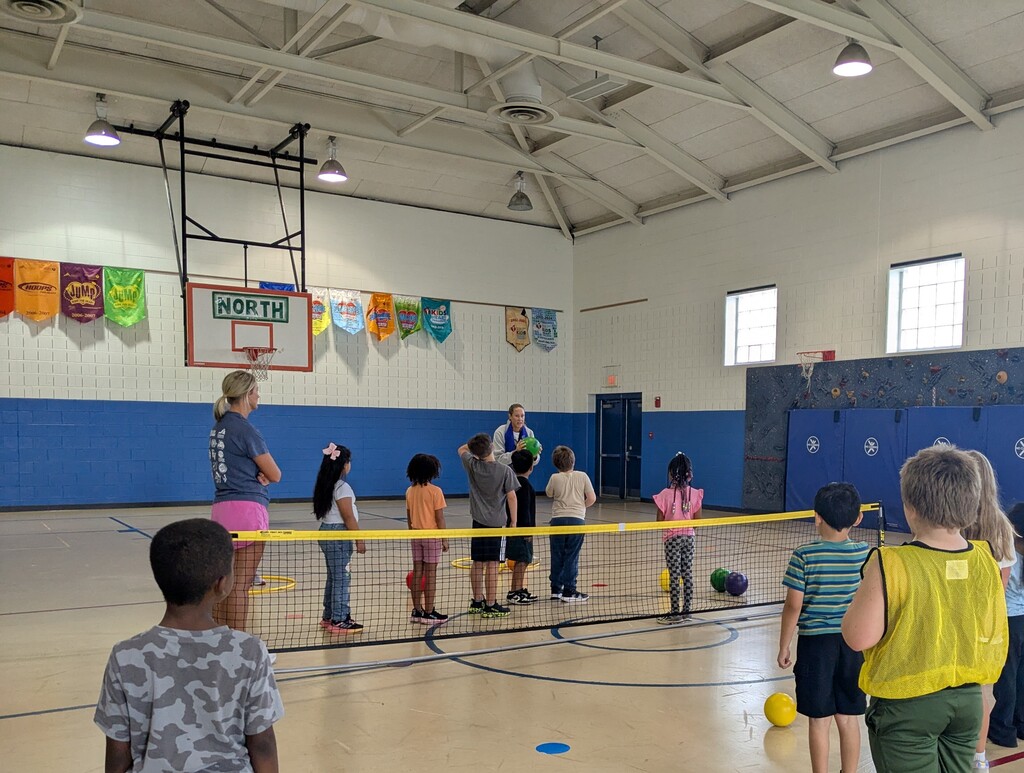 A group of children and adults in a gym with a net and a basketball hoop.