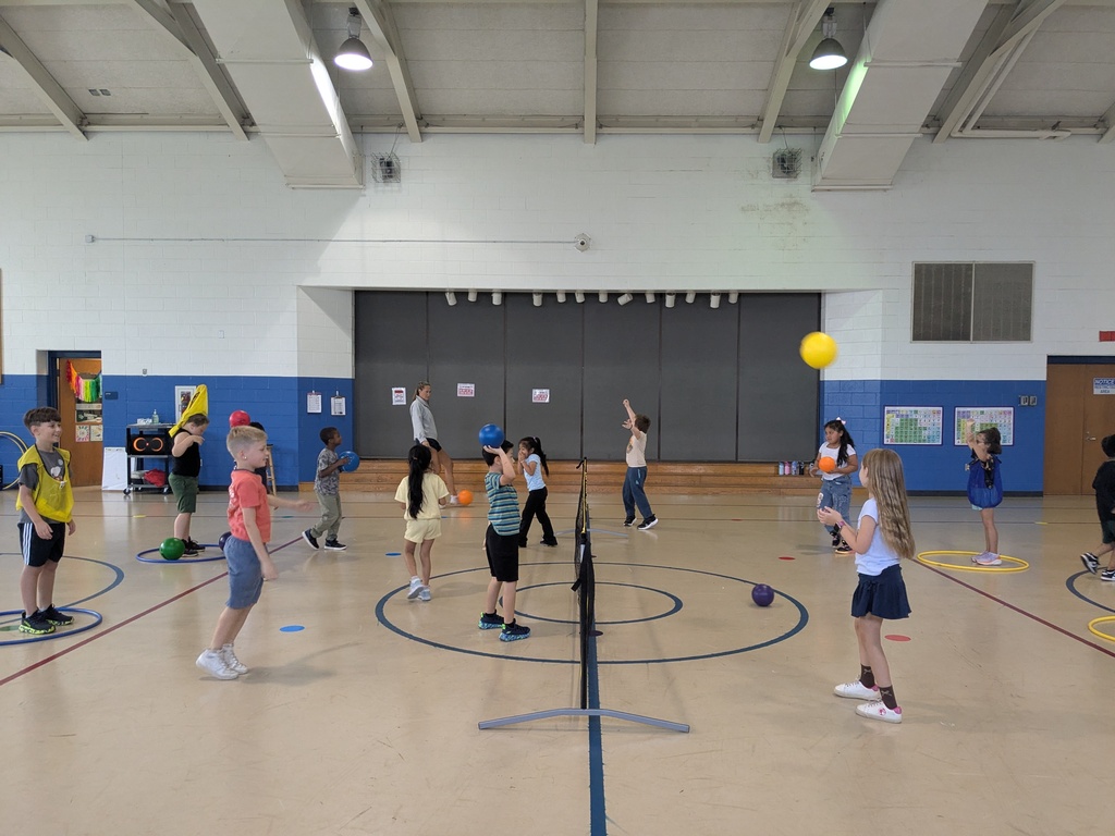 A group of children play a game of ball on an indoor court with blue markings and a yellow ball.