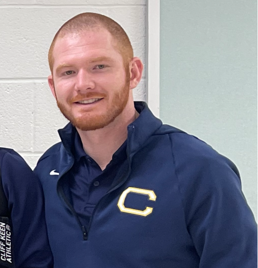 A man with a red beard and mustache smiles, wearing a blue jacket with a yellow "C" on the left side.