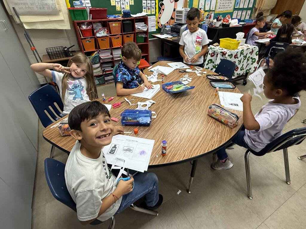 A group of children sit at a round table with various art supplies. Some are engaged in coloring or drawing.
