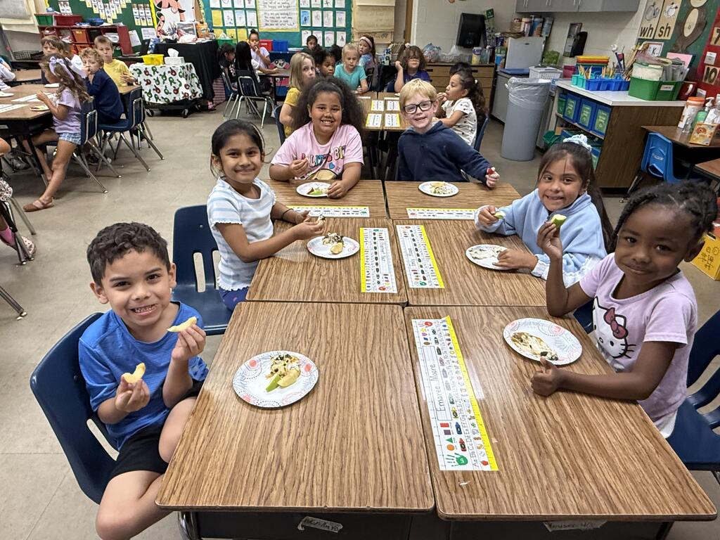 Children sit at tables in a classroom. Some eat sandwiches, and each has a plate. They sit with their backs to the wall.