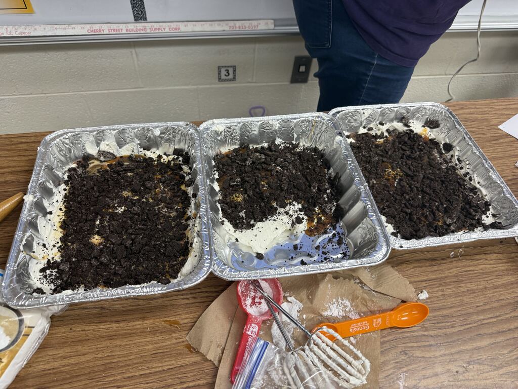 Three foil pans on a table hold soil. One pan has a layer of food waste. A brown bag with utensils is nearby.