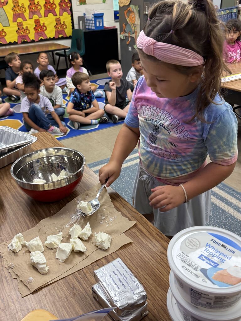 A child in a tie-dye shirt uses a spoon to scoop cheese into a bowl. Other children sit on the floor.