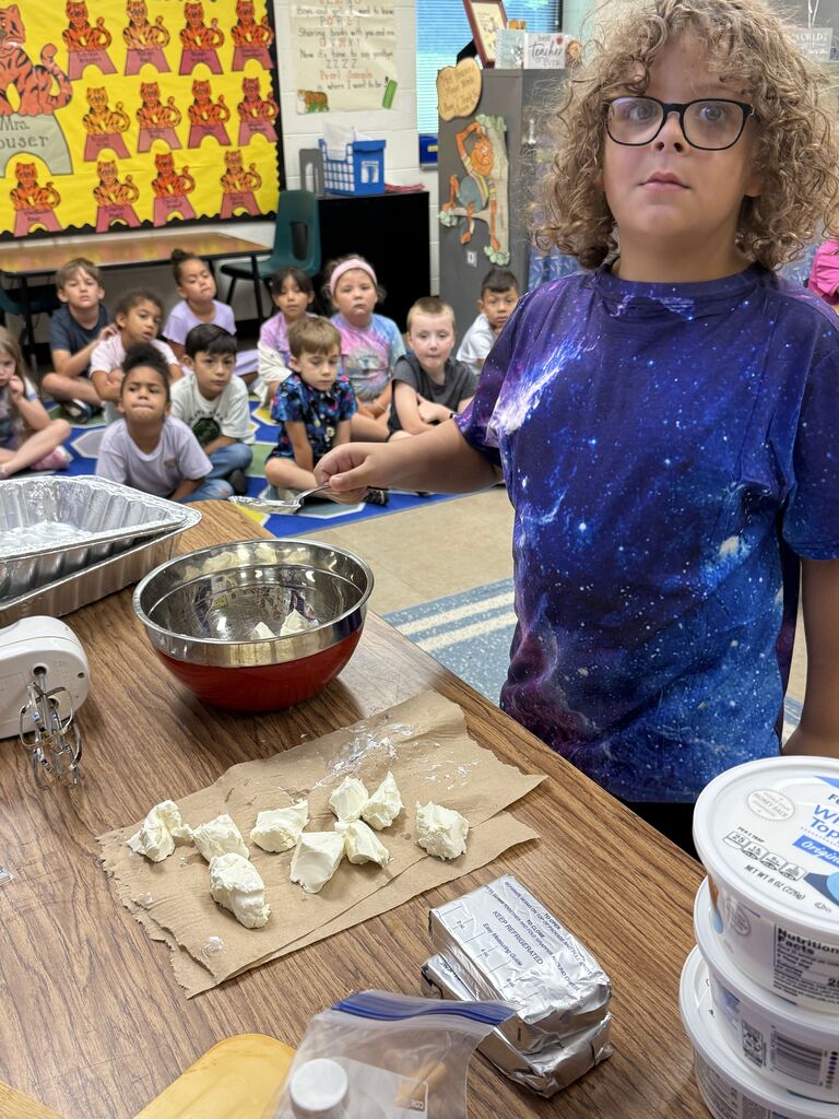 A child in a blue t-shirt prepares food in a classroom. They stand by a table with food items. Children sit on the floor in the background.