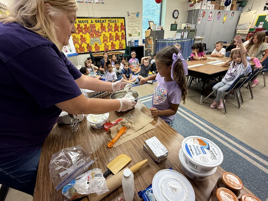A woman guides a young girl at a table with baking ingredients. Other children sit at tables behind them in a classroom.