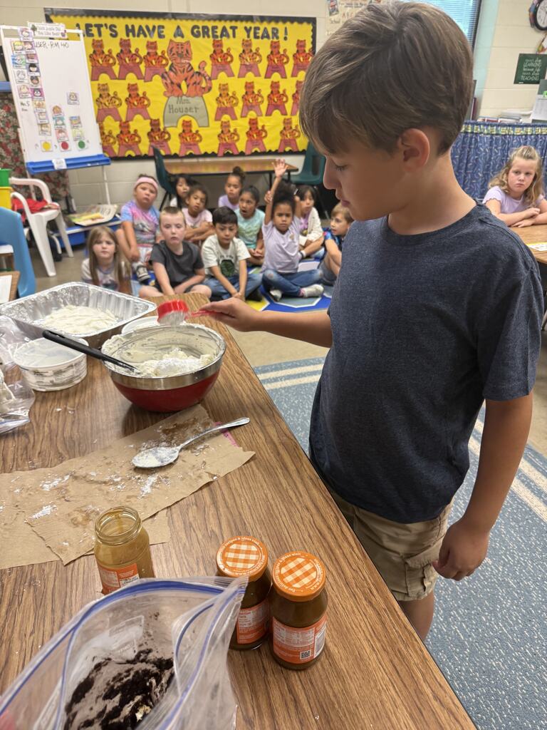 A child holds a spoon, pouring ingredients into a bowl. Behind, children sit on the floor, listening. The table has a bowl, spoon, and jars.