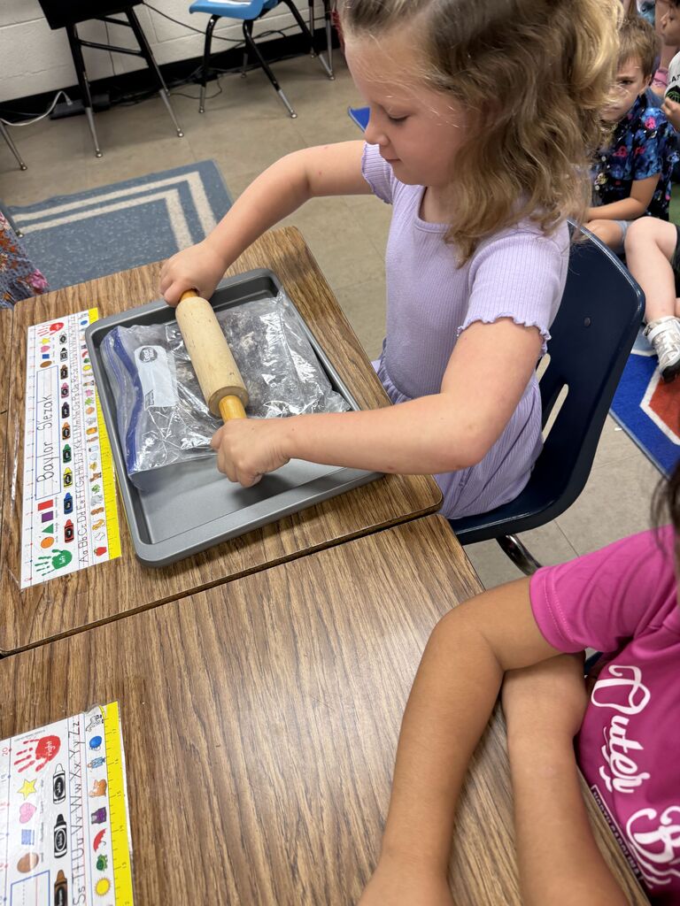 A child rolls dough in a tray at a table in a classroom with another student nearby.