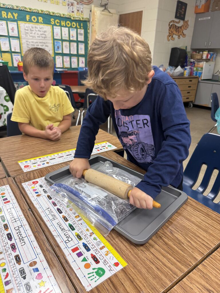 Two children roll dough with a rolling pin on a tray. One wears a yellow shirt; the other wears a blue shirt. Behind them, a classroom setting with desks and chairs.
