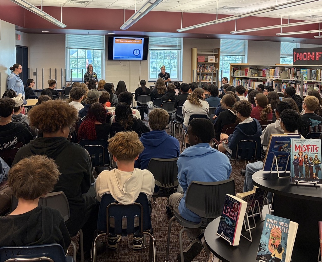 Large group in a library listening to a presentation. Speakers on stage. Books on table.