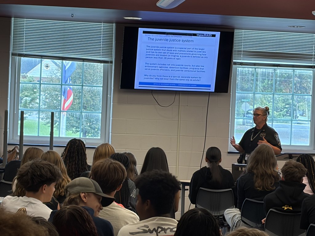 Classroom with a speaker presenting to students. A screen displays information. Students sit in chairs, facing the speaker.