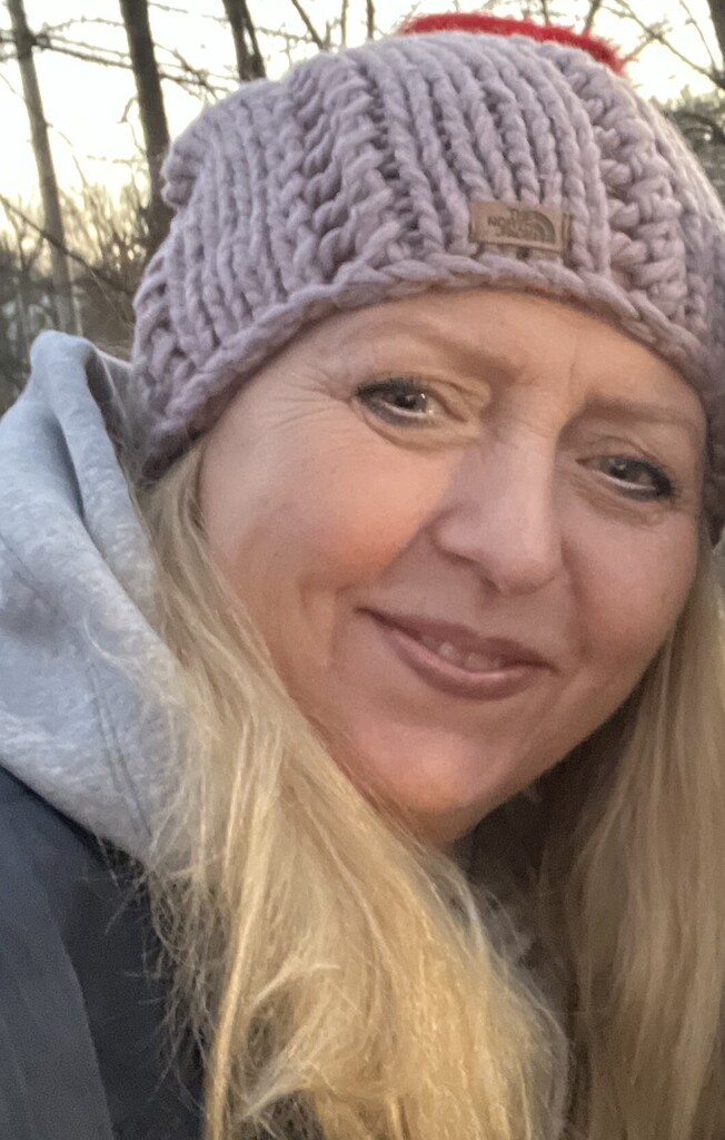 Woman with blond hair, smiling, wearing a knitted hat and gray hoodie against a blurred background.