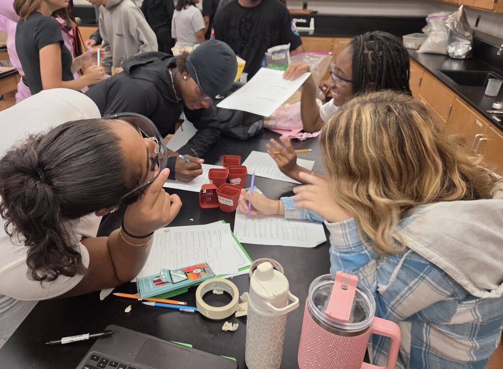 A group of students at a table in a classroom, looking at papers and engaging in discussion.