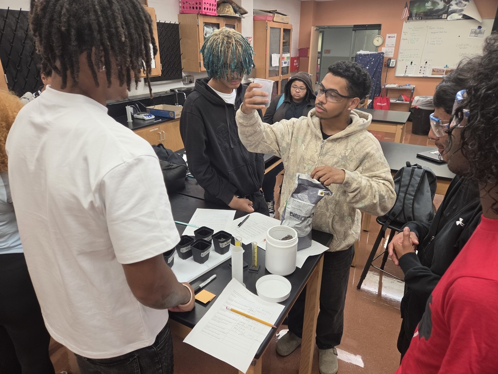 Five students in a classroom. One holds a container. Desk has papers, pens, and a cup.