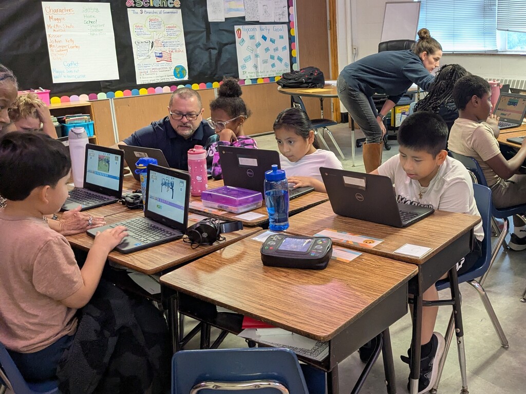 Students sit at desks with laptops. A teacher stands behind a student. White papers are on the bulletin board.