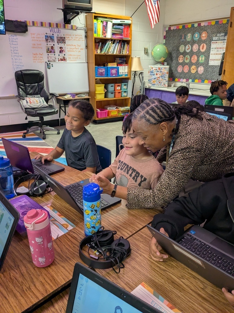 A woman and two children are using laptops at a table. Behind them is a classroom with bookshelves and a whiteboard.