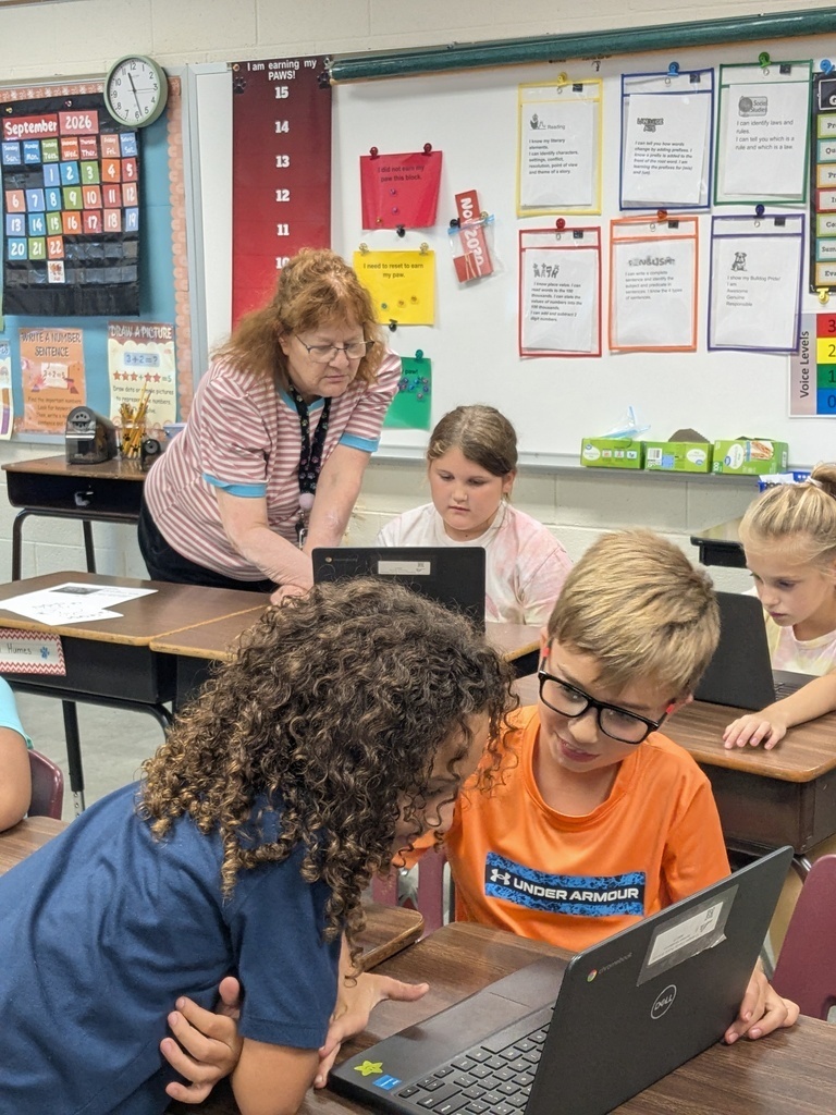 A teacher and several children use laptops at desks in a classroom. Papers and clocks are visible.